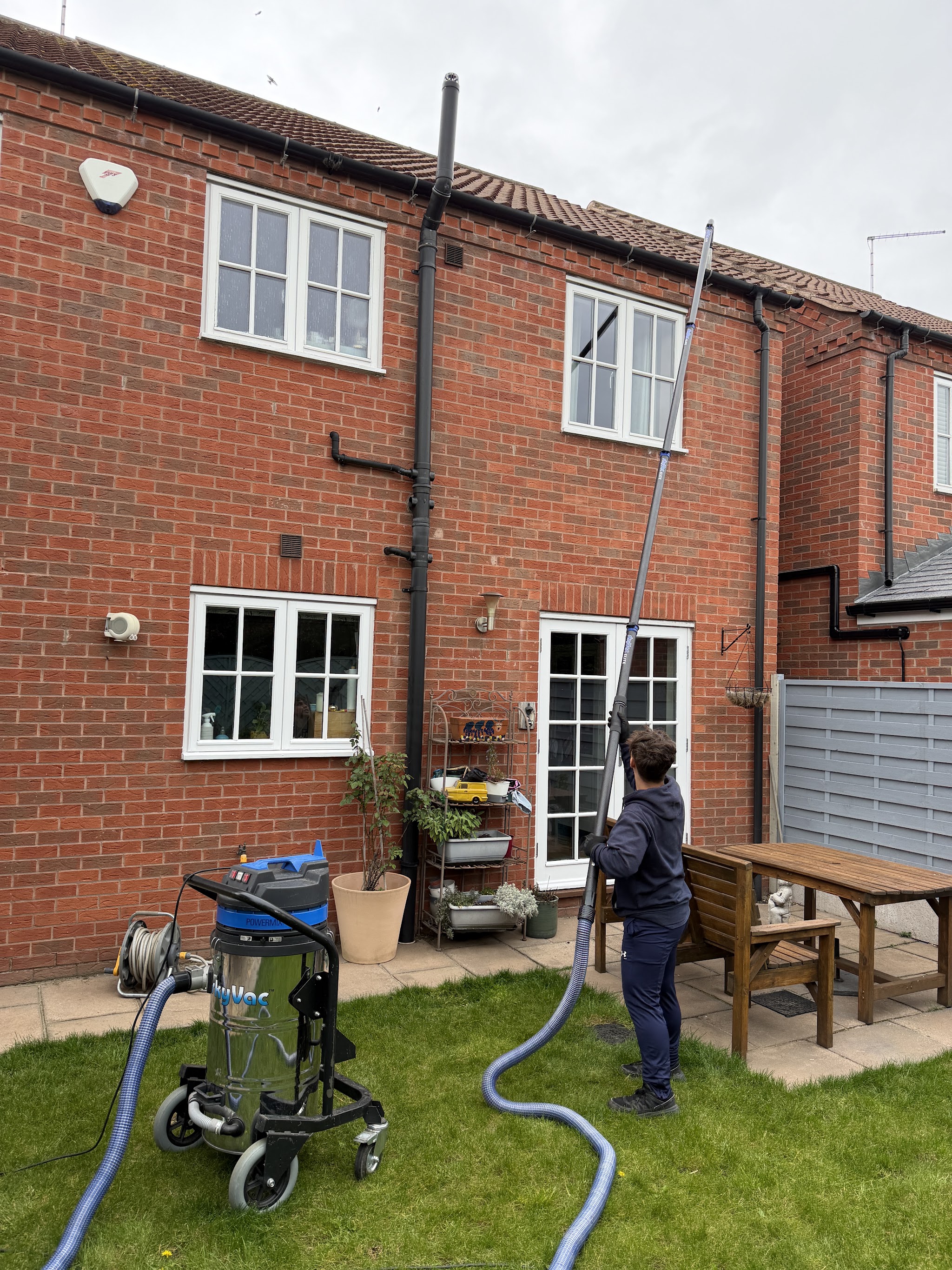 A Purereach cleaner washing the upstairs windows of a brick house from the back garden