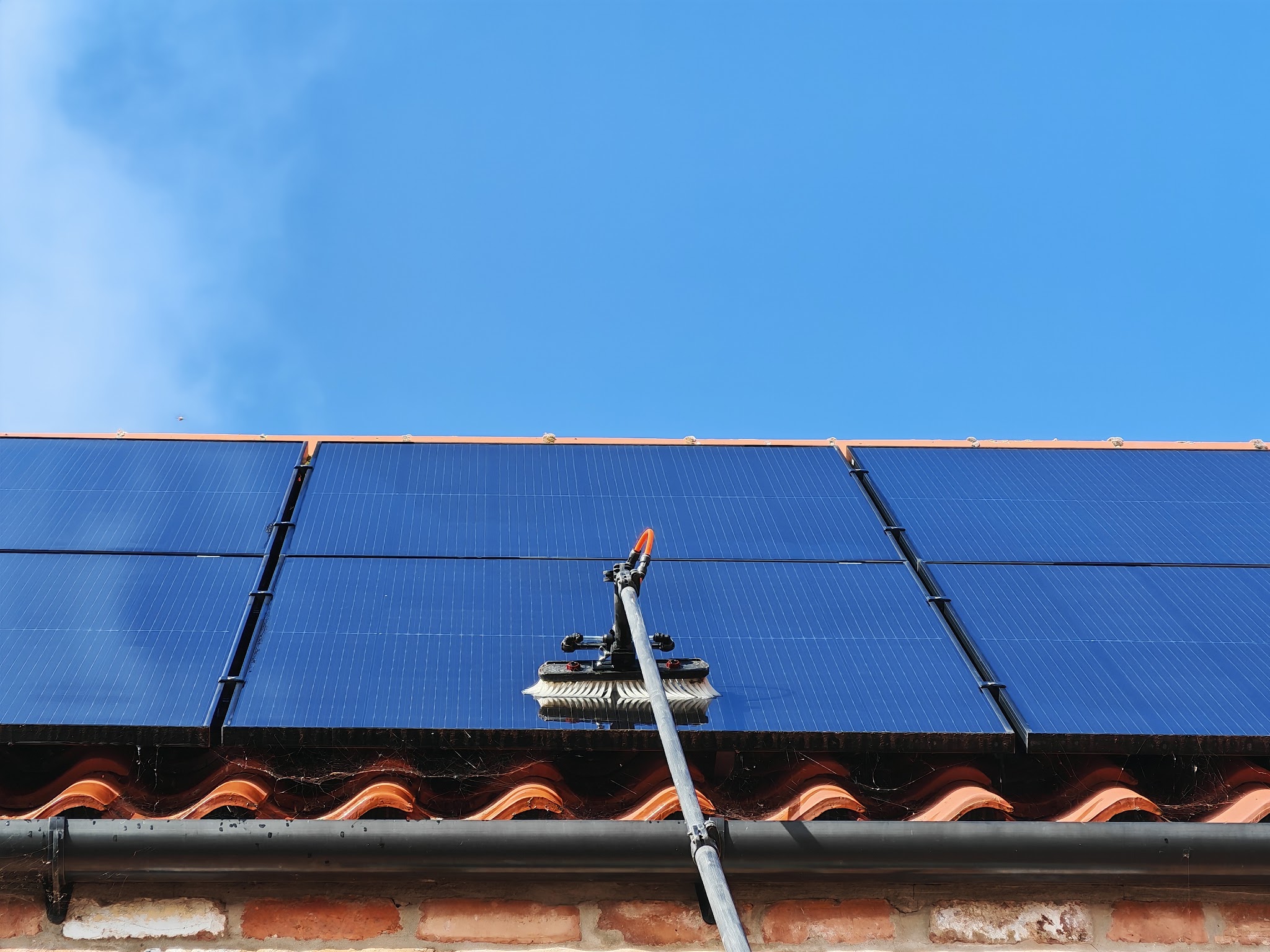 A brush on a pole cleaning a row of roof solar panels under a clear blue sky